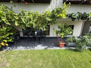 An outdoor patio with a table and chairs, surrounded by greenery and a white wall.