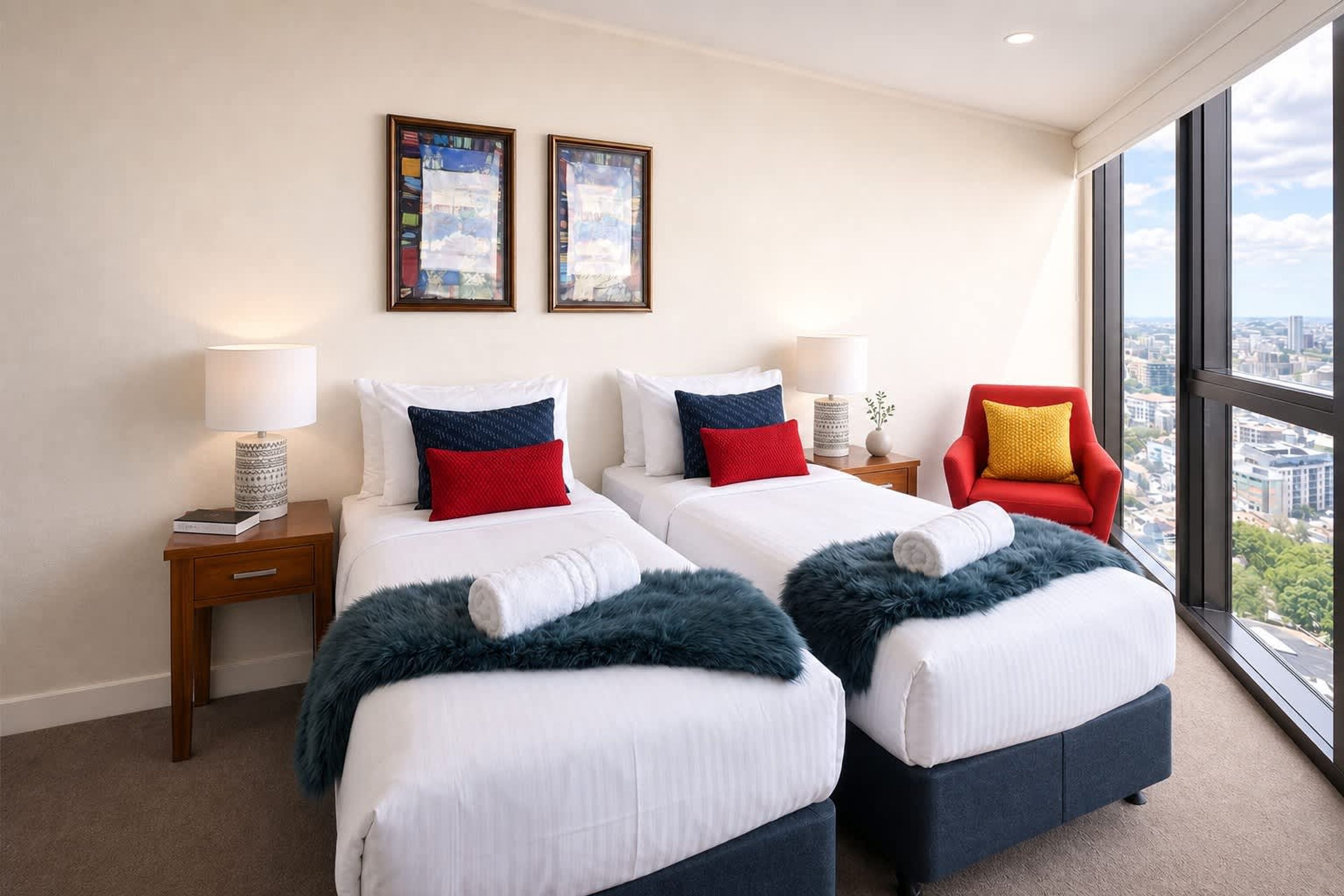 A hotel room with two beds, a red chair, a large window, and two framed pictures on the wall.