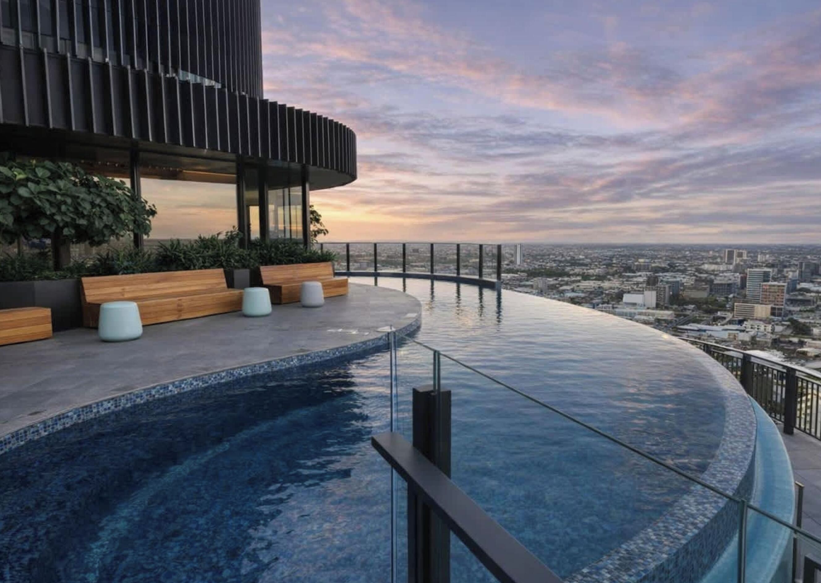 Una piscina infinita en la azotea con vista a la ciudad, mobiliario de madera y una estructura de edificio moderna.
