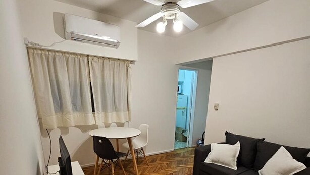A compact living space with a ceiling fan, a white air conditioning unit, a black sofa with white cushions, a white desk with a computer, and a herringbone-patterned wooden floor. - Comfortable and Well-Located in Recoleta (Buenos Aires)