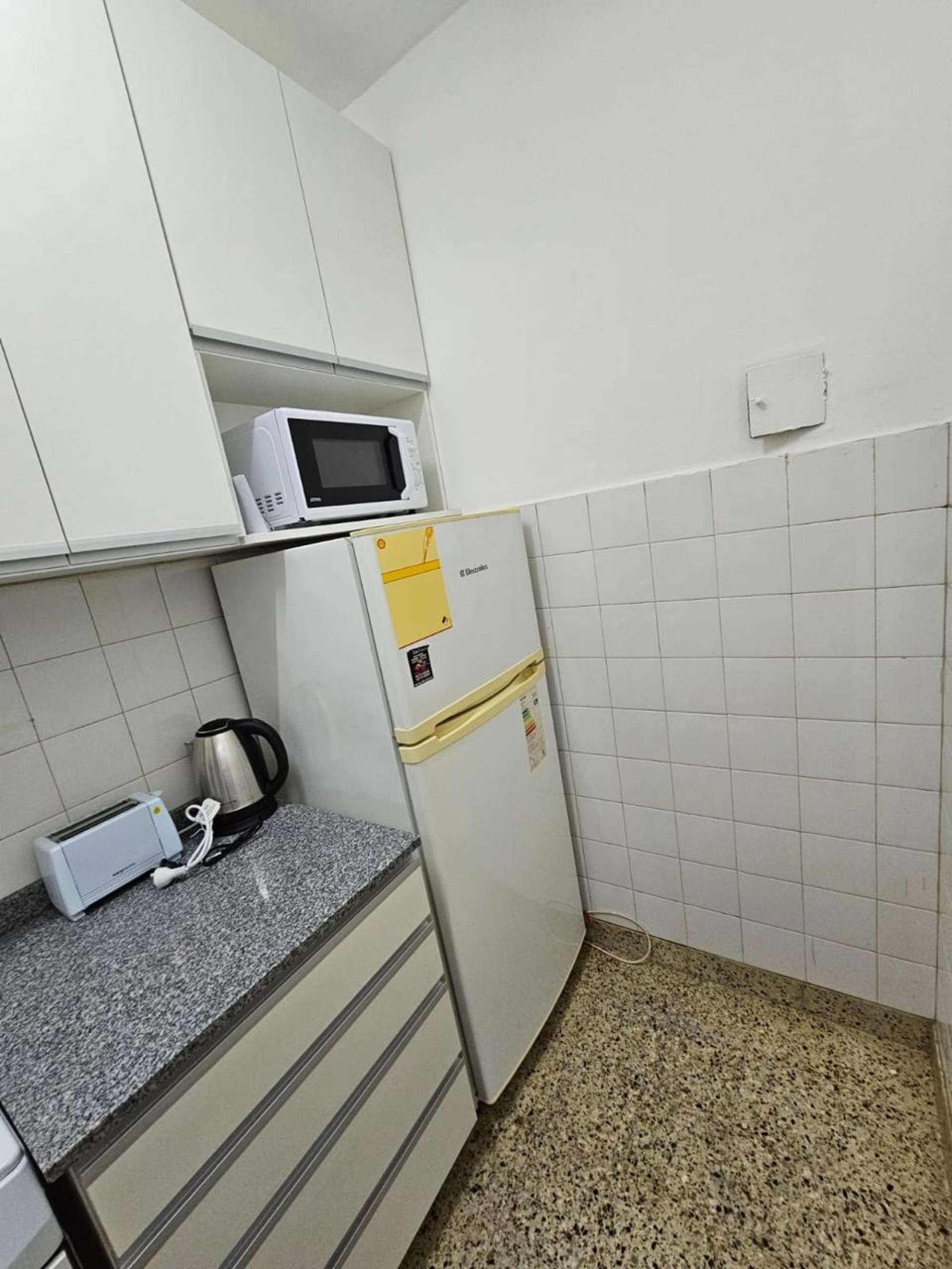 Une cuisine compacte avec des meubles blancs, un four à micro-ondes, un petit réfrigérateur, une bouilloire et un grille-pain posés sur le plan de travail.