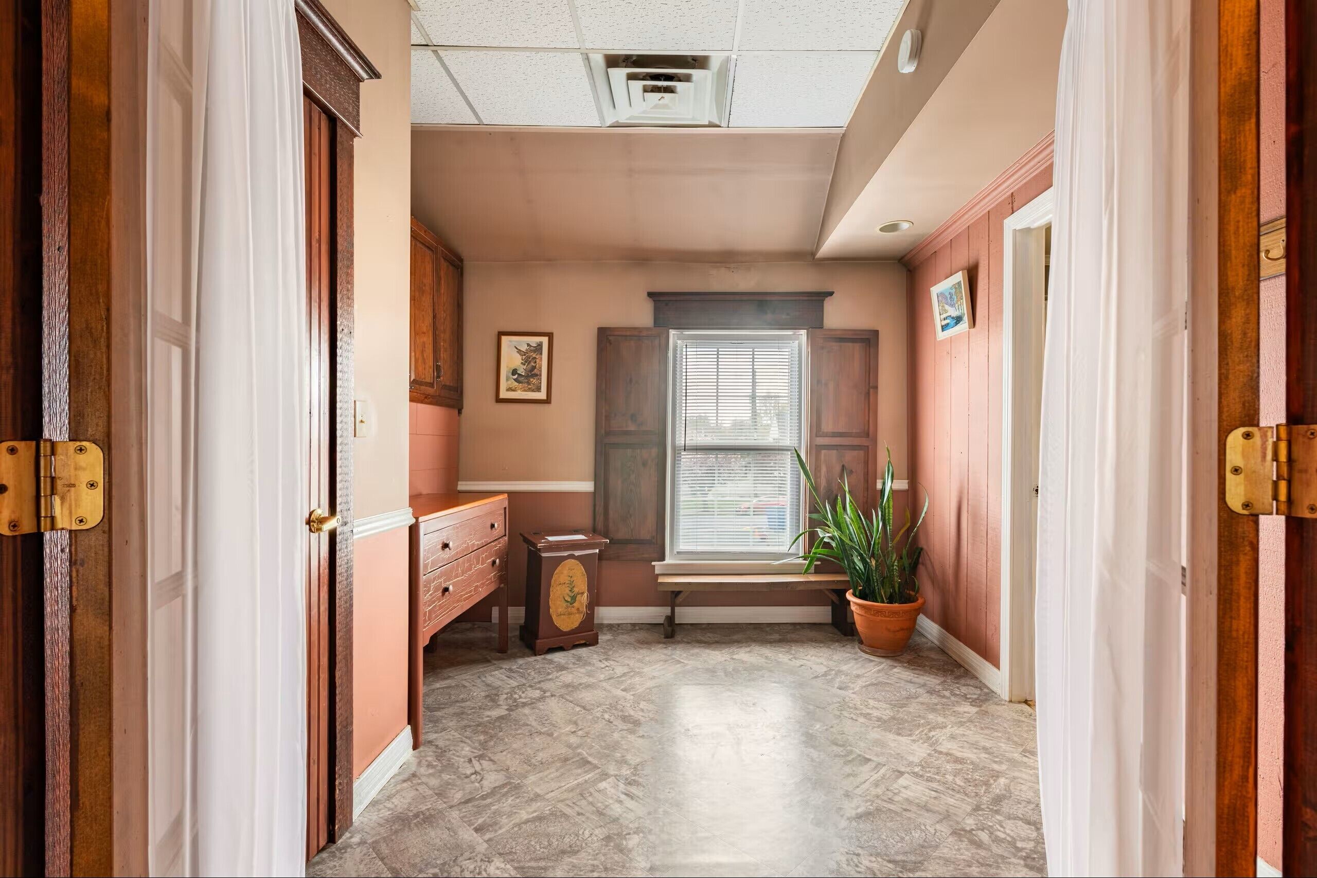 A hallway with a bench, a window with blinds, a potted plant, and a door with a latch.