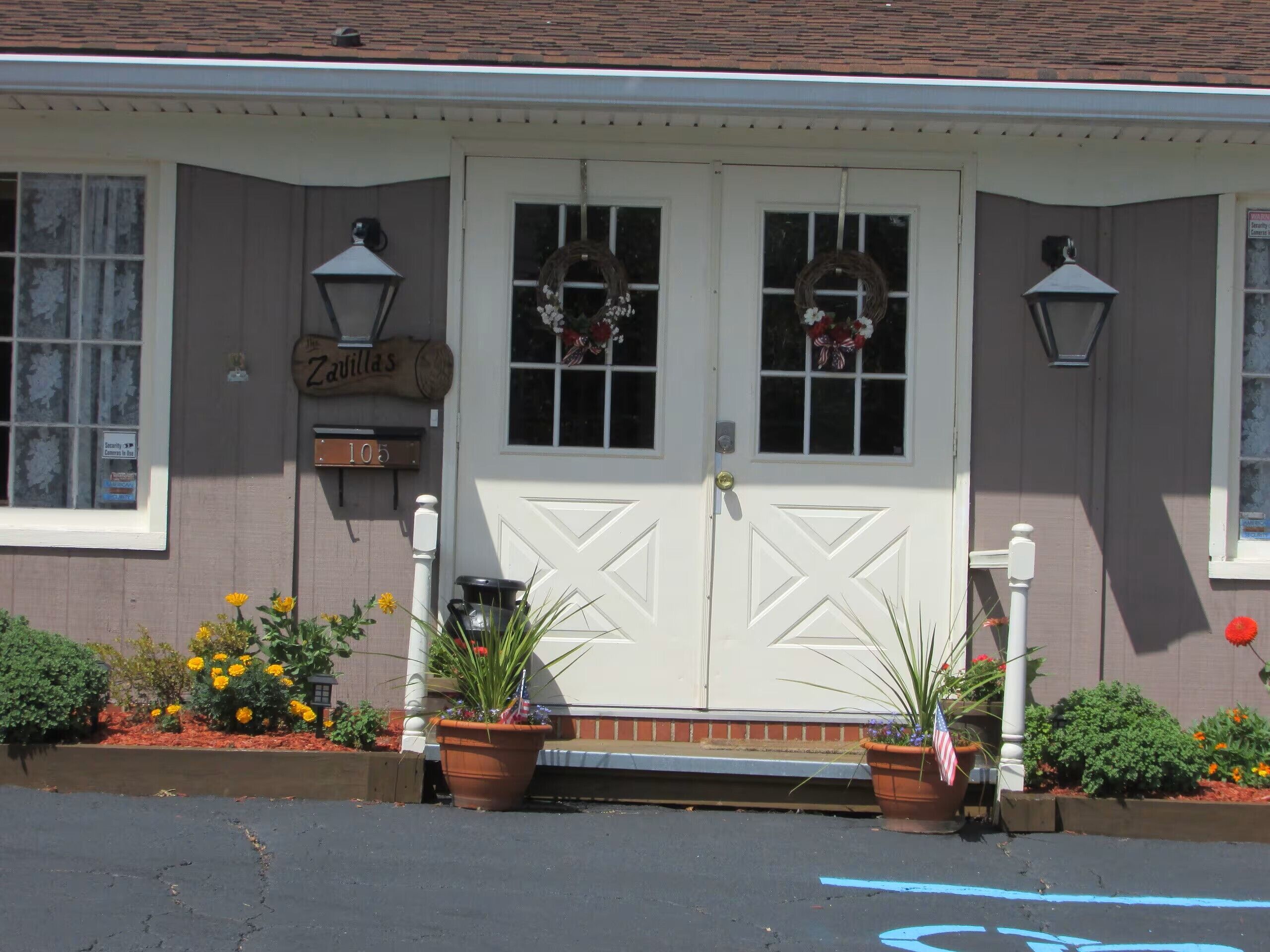 A building with a white front door, two windows with wreaths, a sign above the door, and potted plants in front.