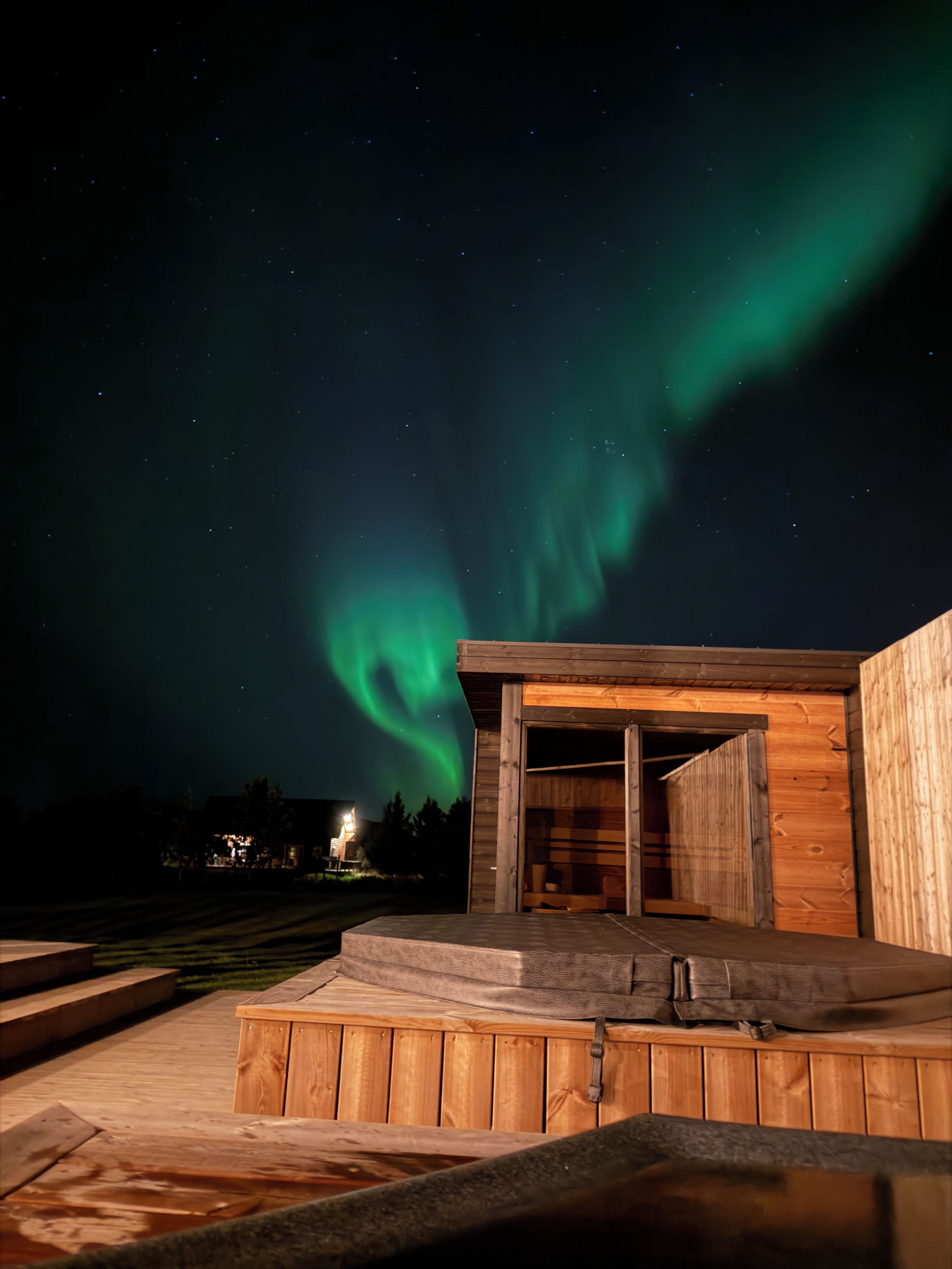 A wooden cabin with an open door, a hot tub, and a view of the northern lights.