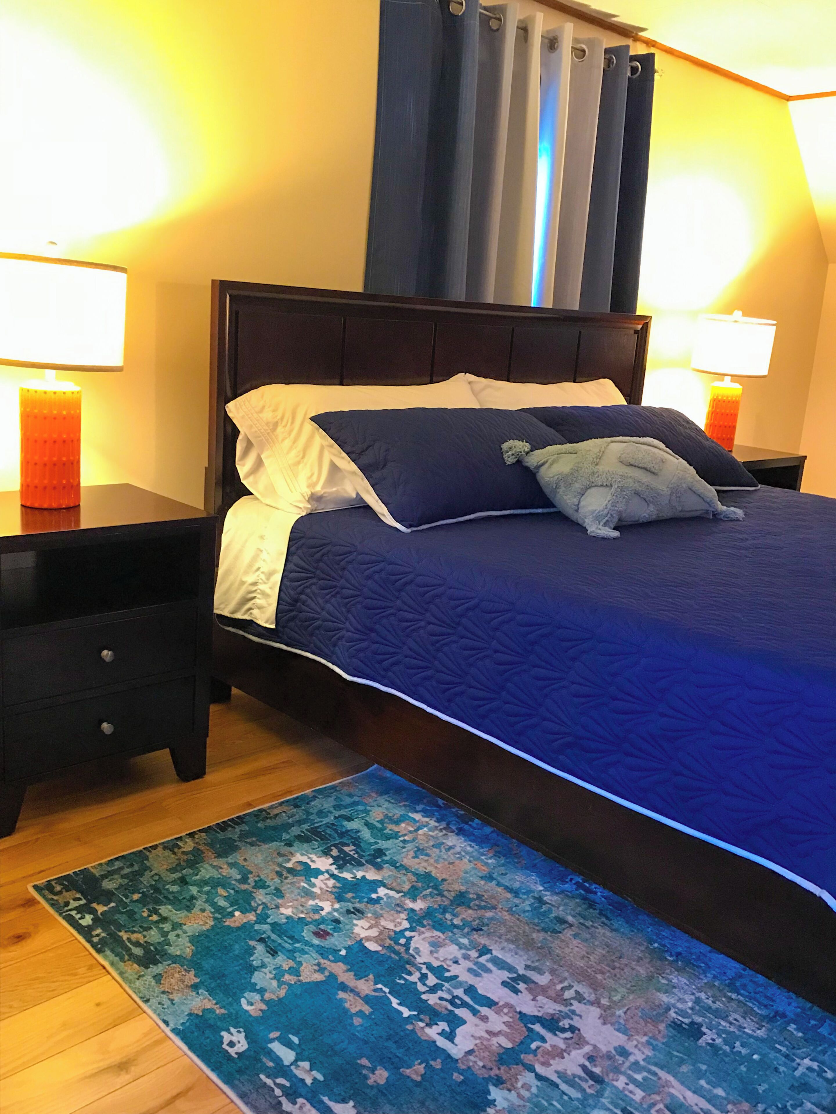 Un lit avec une tête de lit sombre, deux tables de chevet avec des lampes, un tapis à motifs bleus et une fenêtre avec des rideaux.