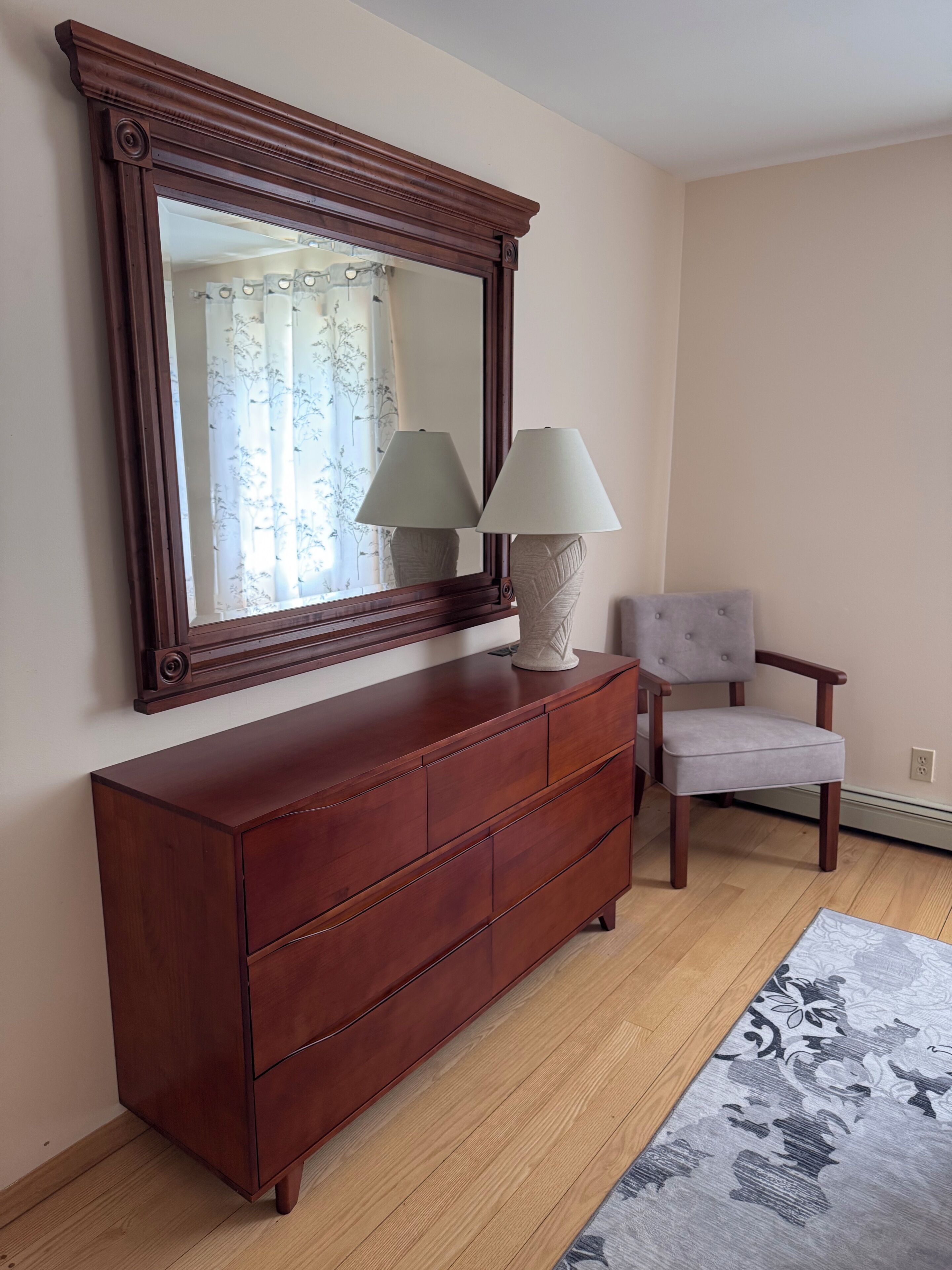 Un aparador de madera con dos lámparas, un espejo y una silla en una habitación con piso de madera y un tapete estampado.