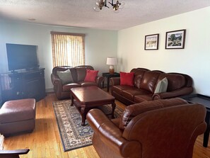 A living room with a brown leather sofa, a wooden coffee table, a TV on a stand, and a window with blinds.