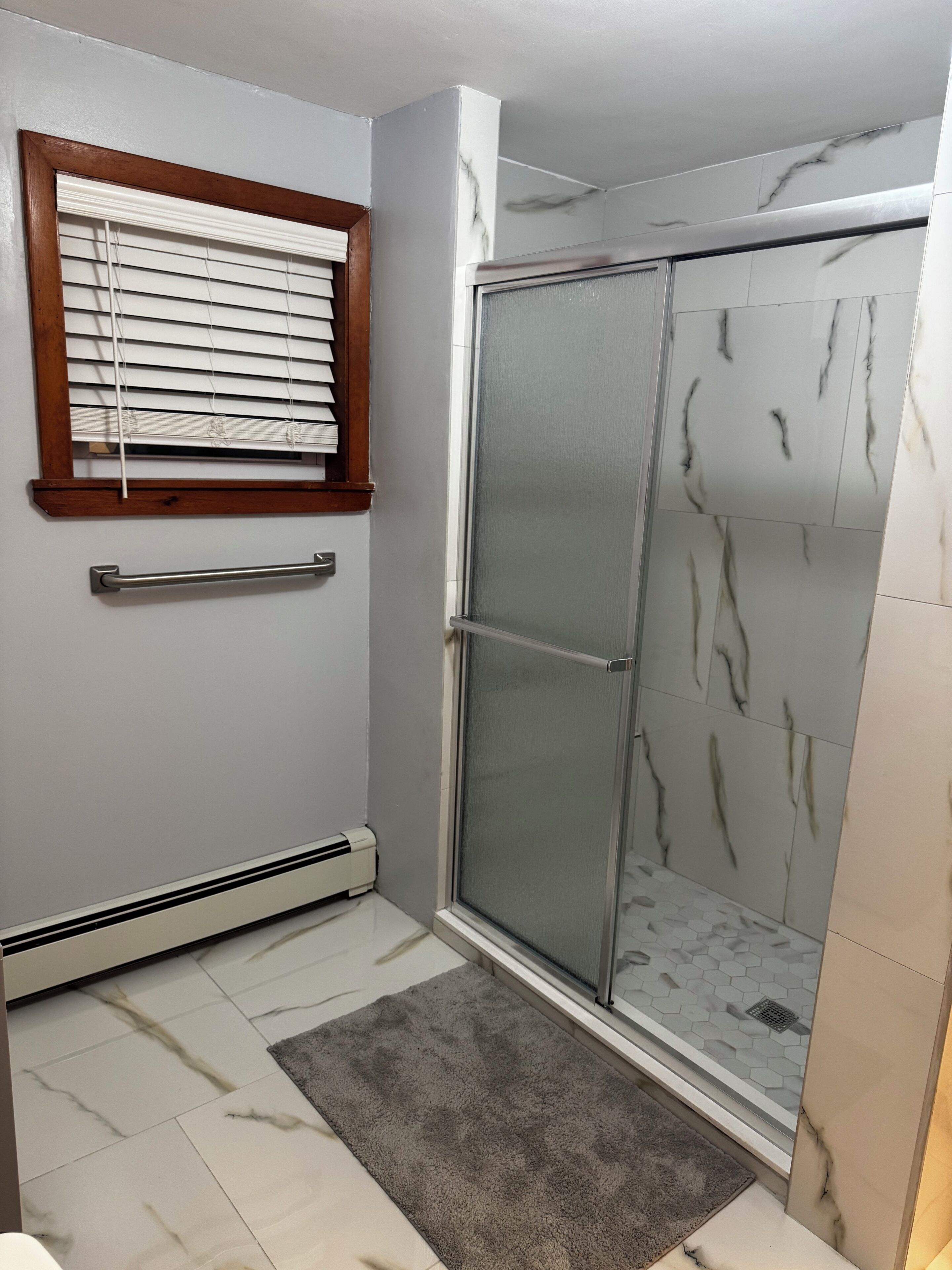 A shower area with a glass door, a towel rack, and a window with blinds.