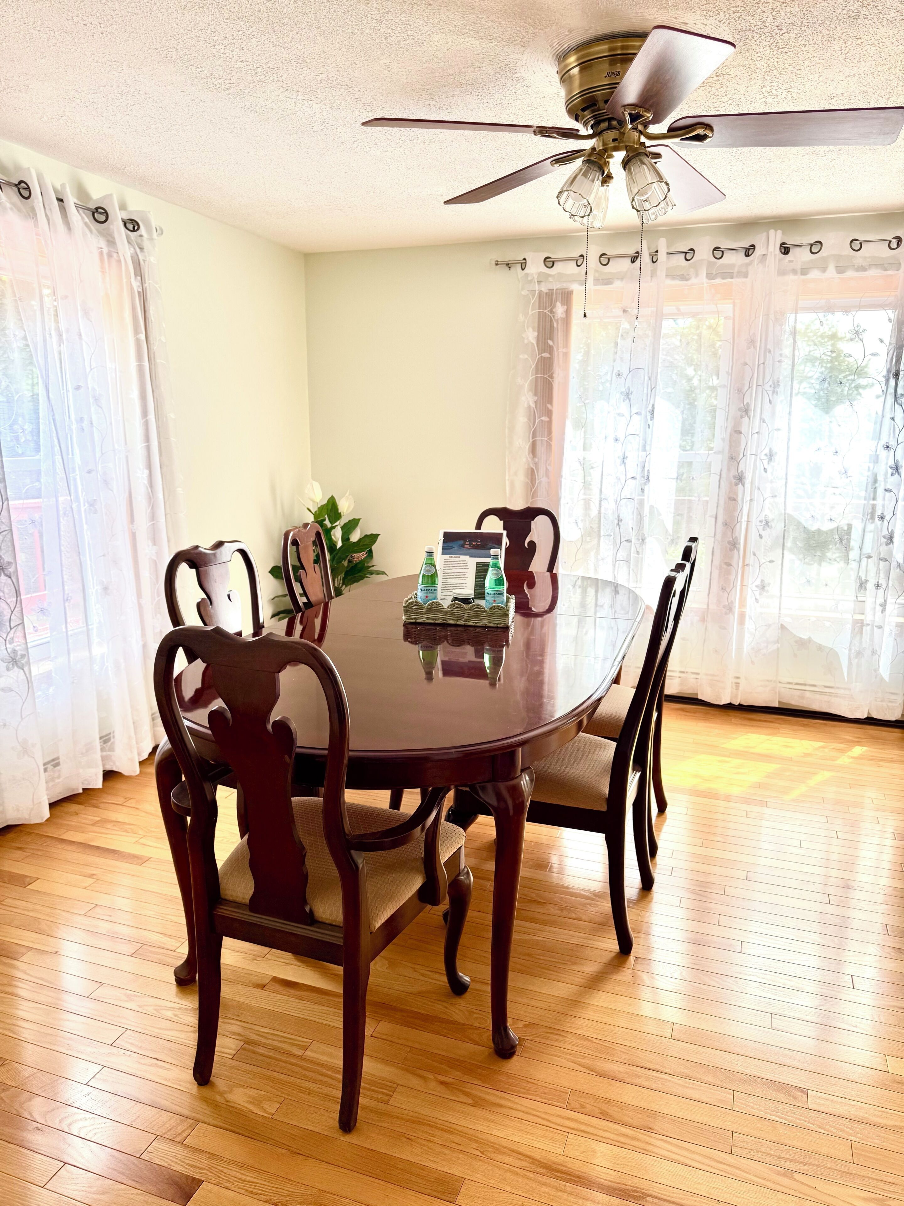 A dining area with a round table, wooden chairs, a ceiling fan, and a window with curtains.