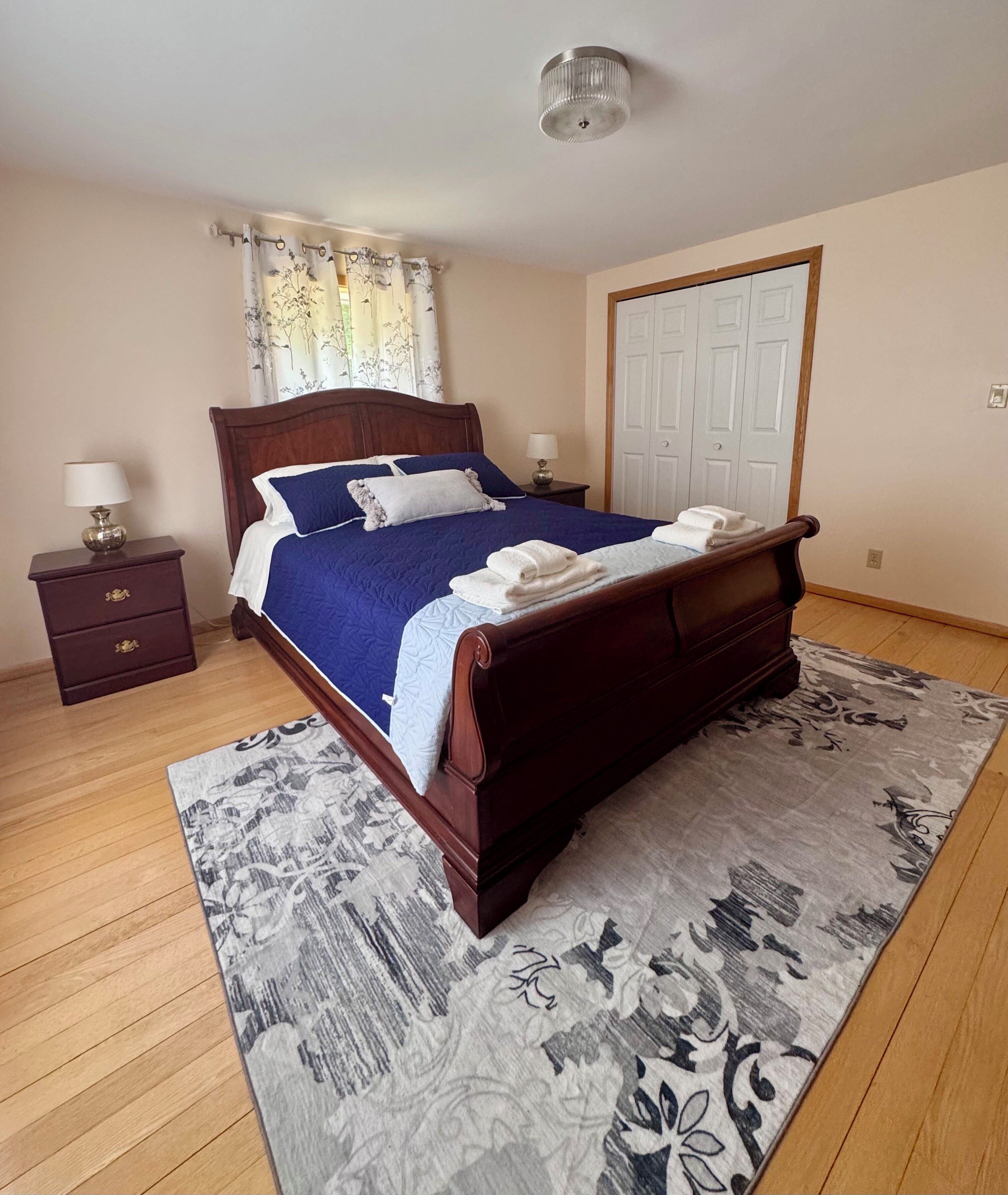 A bedroom with a wooden bed, a nightstand, a lamp, a window with curtains, and a door.