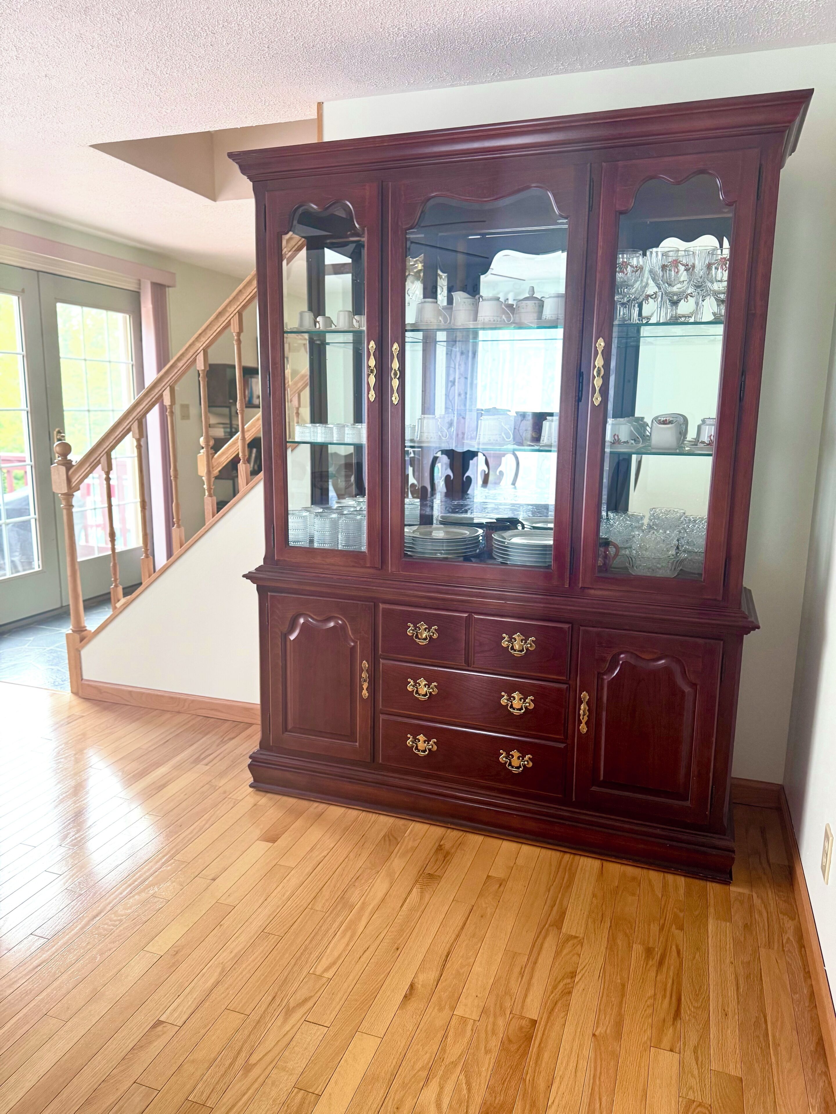 A wooden cabinet with glass doors and drawers, located in a room with wooden flooring and a staircase.