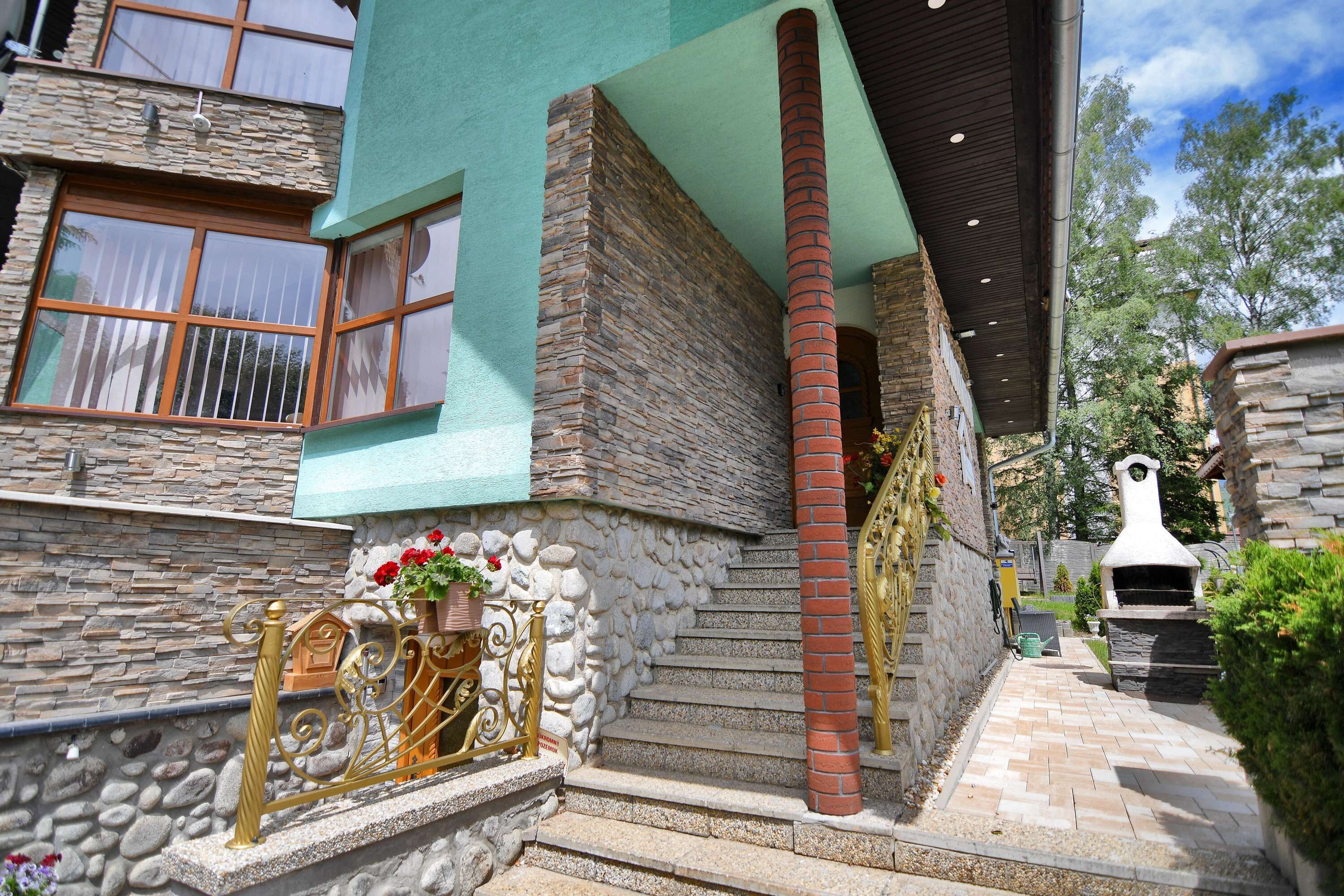 Un bâtiment en pierre avec une façade verte, une cheminée en briques et un petit espace extérieur doté d’un barbecue et d’un escalier menant à une porte.