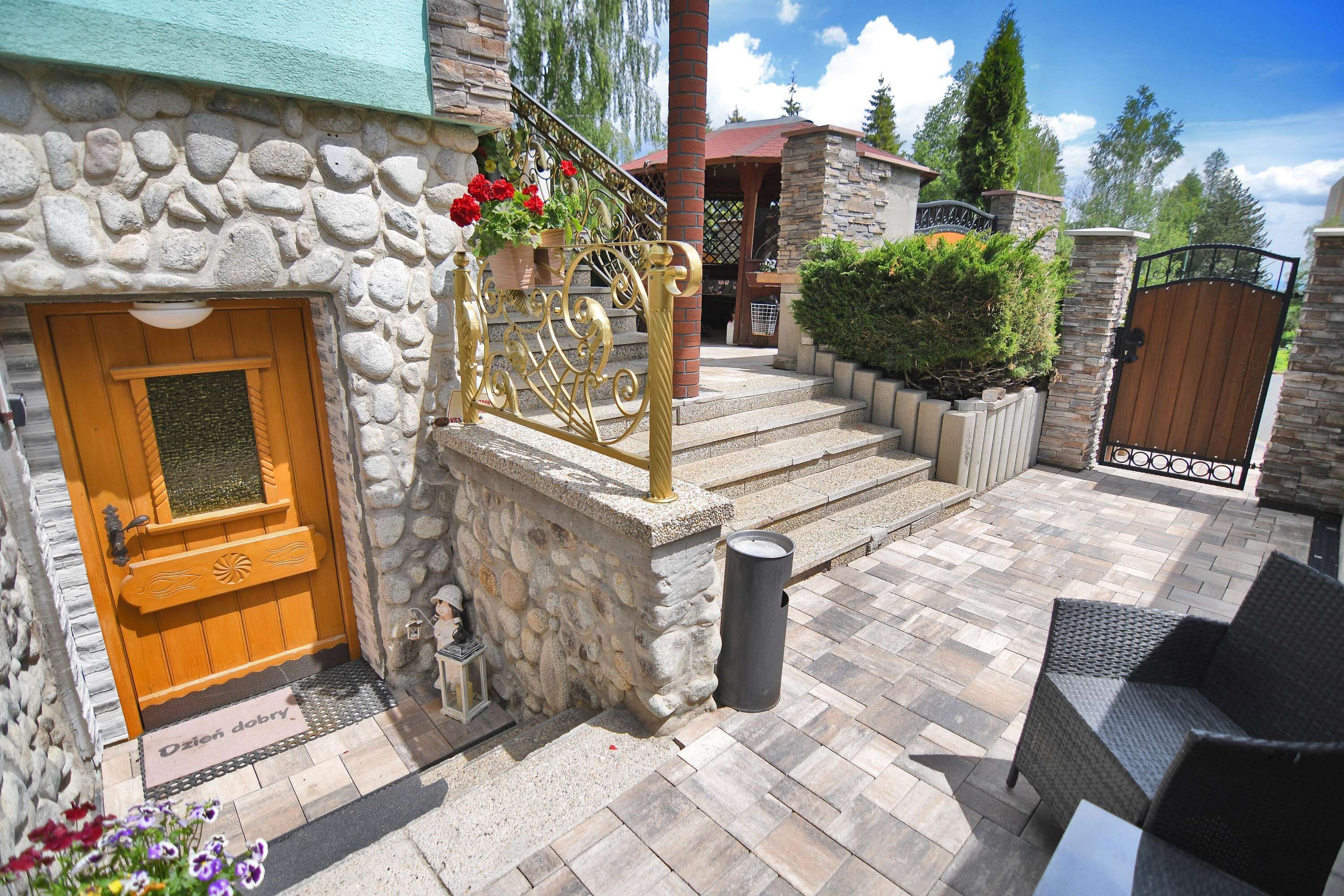 Un bâtiment en pierre avec une porte en bois, un portail en métal et une terrasse dallée aménagée avec du mobilier d’extérieur.