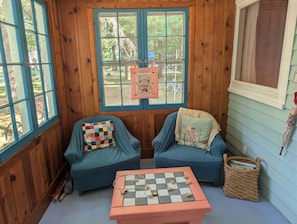 A cozy room with two blue armchairs, a checkered cushion, a small table with a chessboard, and a window with a view of trees. - Cozy 1-bedroom/ 1 loft cottage in serene Lakeview- sleeps 8 (Lakeview)