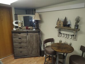 Una cucina compatta con un pensile in legno, un piccolo tavolo da pranzo con due sedie e uno scaffale a parete con oggetti decorativi.