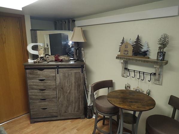A compact kitchen with a wooden cabinet, a small dining table with two chairs, and a wall-mounted shelf with decorative items.