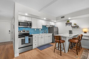 Eine moderne Küche mit weißen Schränken, einem Edelstahlbackofen und einer blauen Spritzschutzwand. Eine kleine Kücheninsel mit Hockern und einer Kaffeemaschine steht ebenfalls dort.
