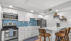 A modern kitchen with white cabinets, a stainless steel stove, a microwave, and a refrigerator. There are two wooden chairs at a small island table. - NEW! STEPS from the Beach and Pool! Beautiful Ocean View 2nd Floor Studio! (New Smyrna Beach)