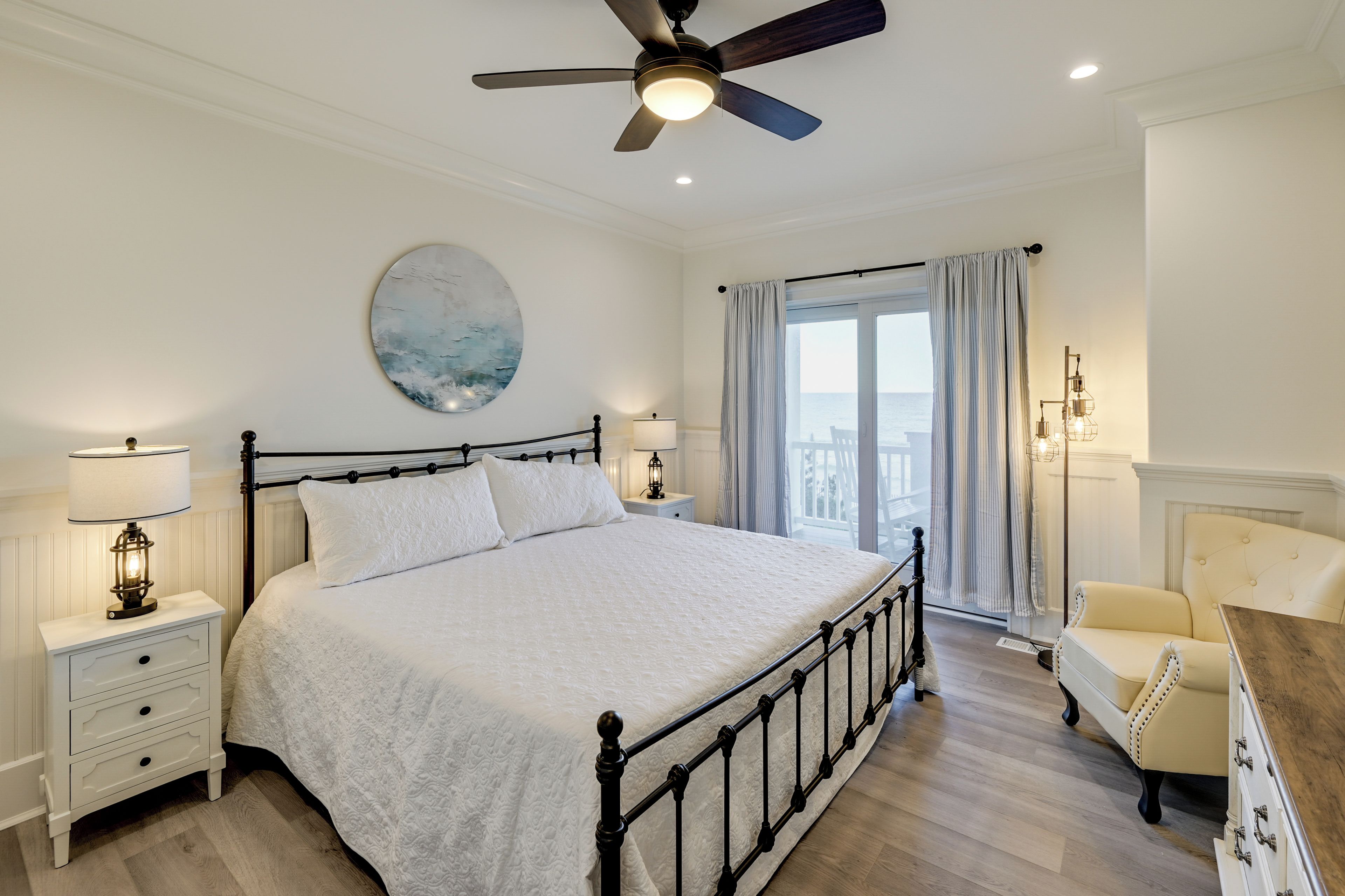 A bedroom with a bed, nightstands, a ceiling fan, a painting, a window with curtains, and a chair.
