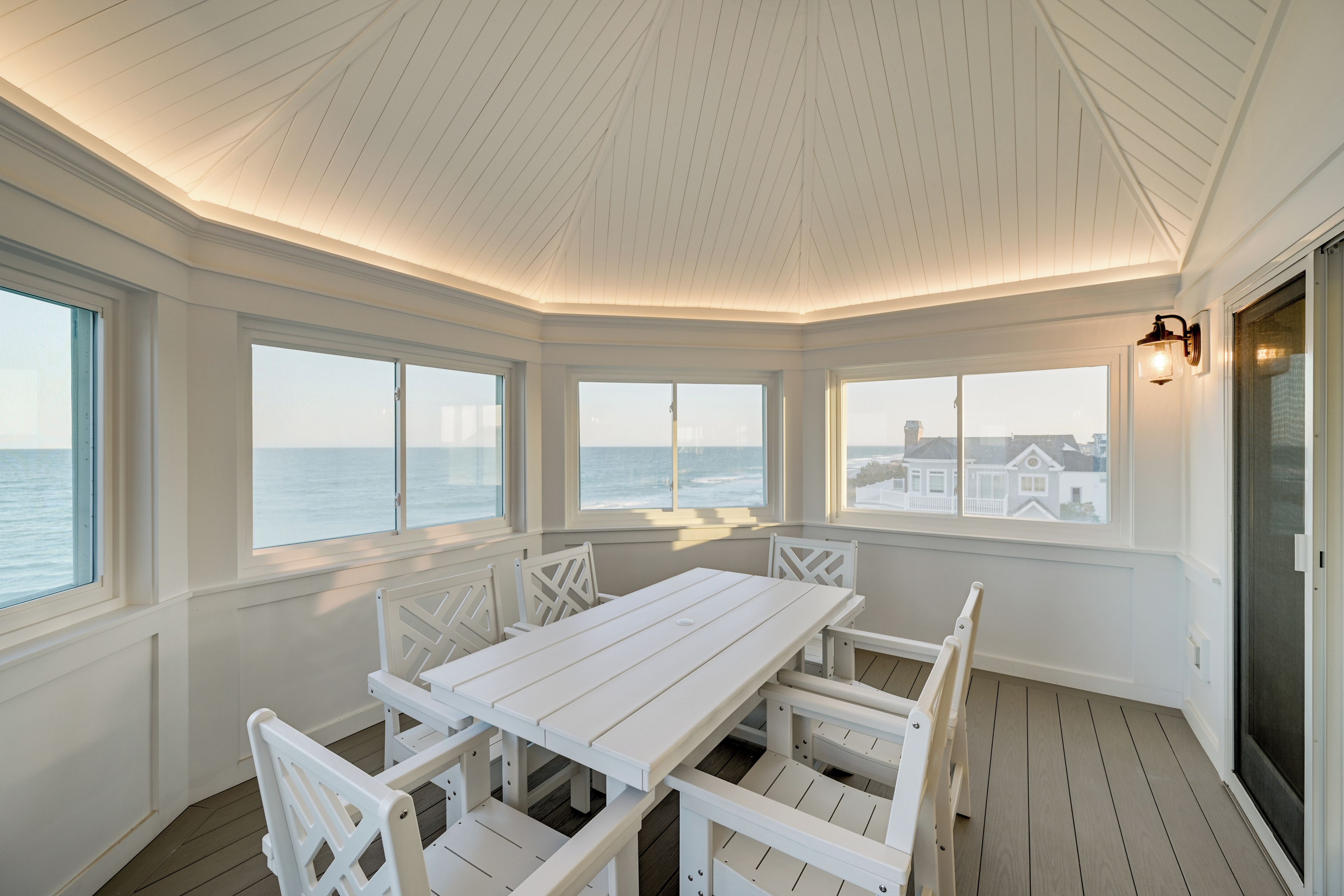 Quarto com mesa e cadeiras brancas, grandes janelas e vista para o mar.
