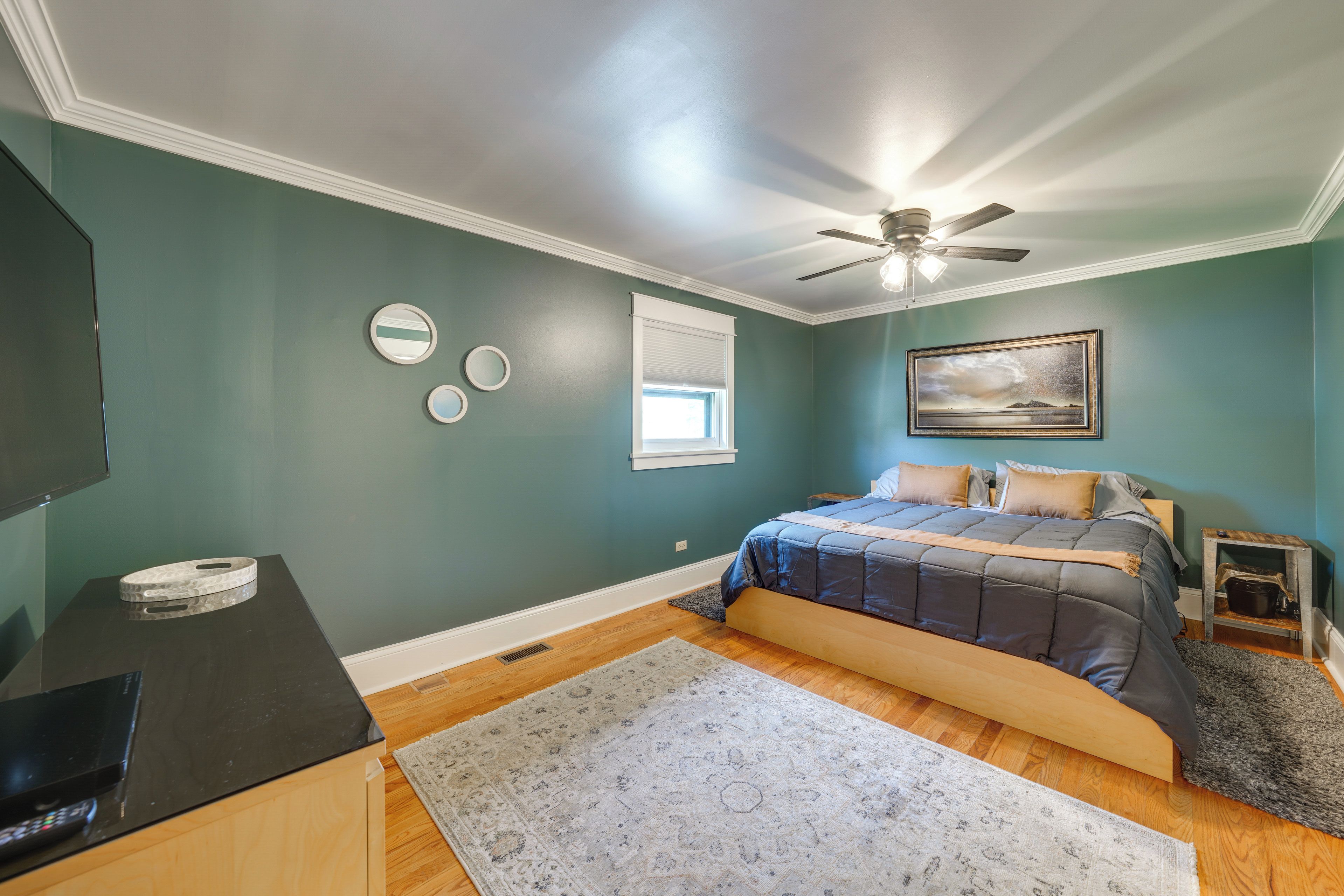 A bedroom with a bed, a ceiling fan, a window, and a painting on the wall.