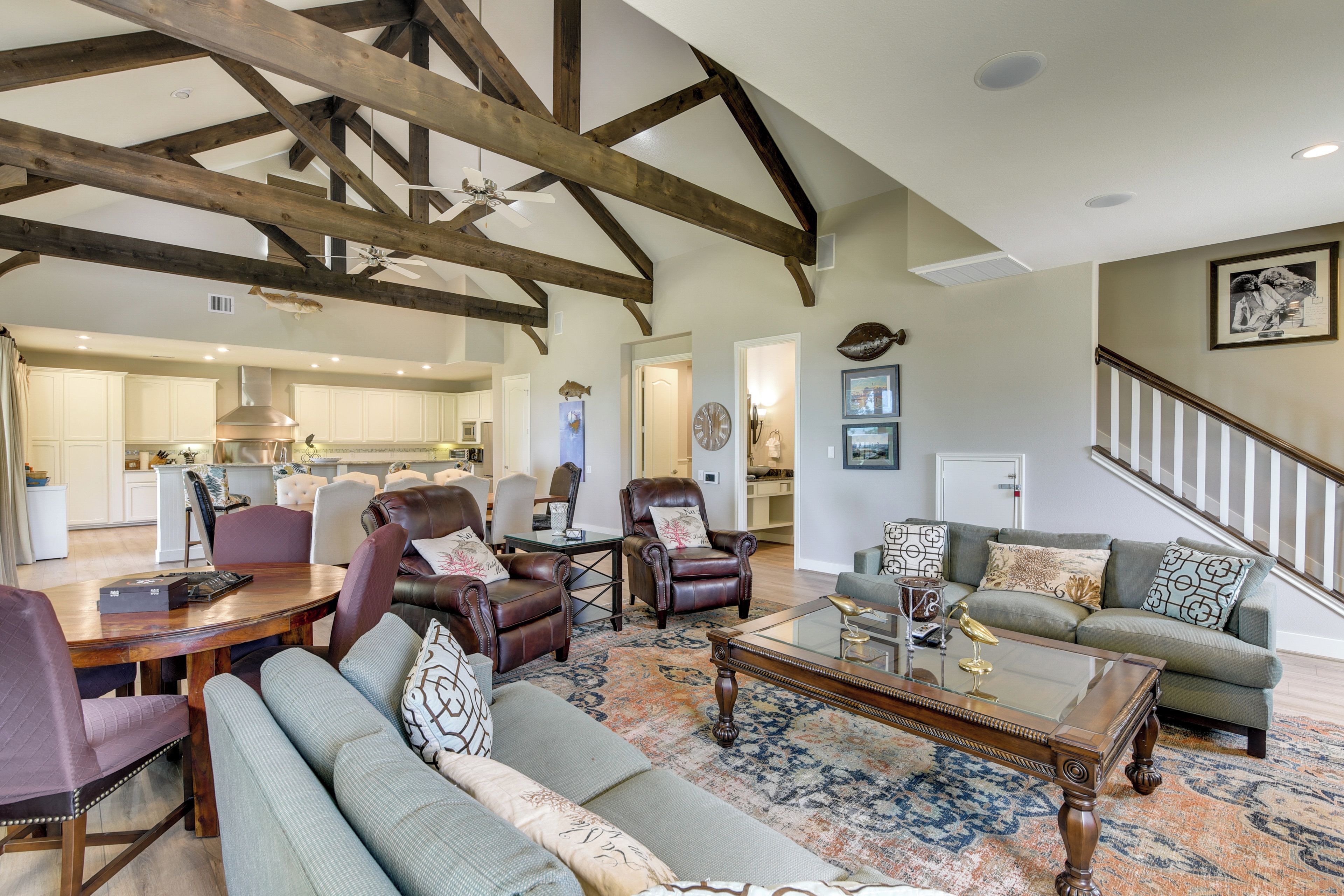 A spacious living room with a high ceiling, exposed wooden beams, a dining area with a table and chairs, and a kitchen in the background.