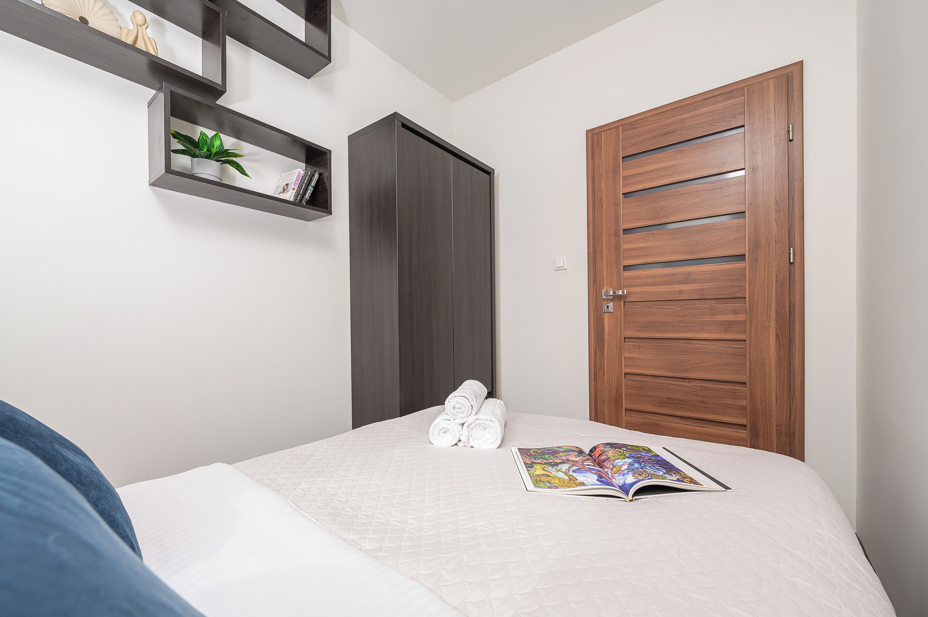 A bedroom with a bed, a wooden door, a shelf with a plant, and a magazine on the bed.