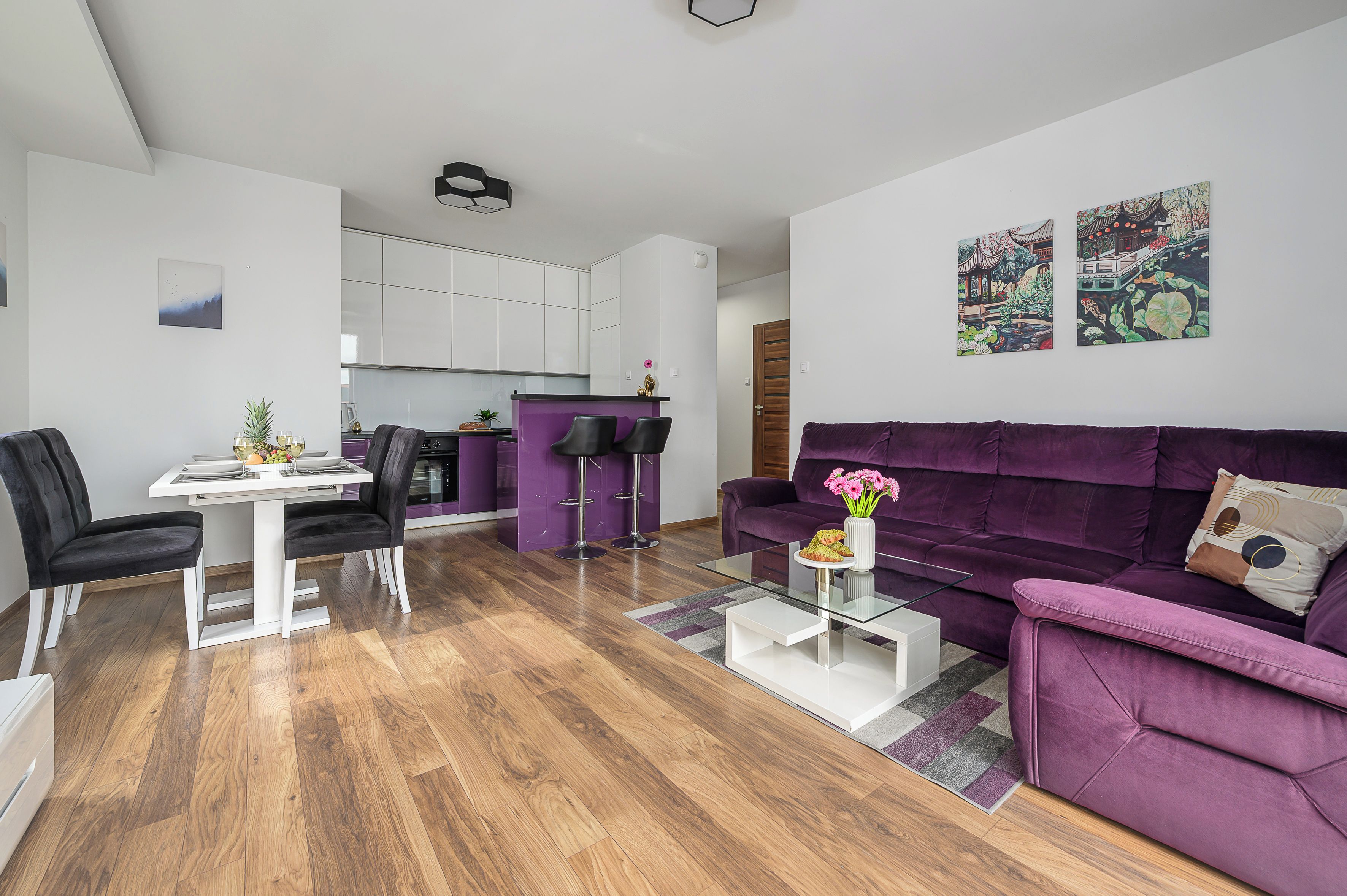 A modern living space with a purple sofa, a dining area with a white table, and a kitchen with white cabinets.