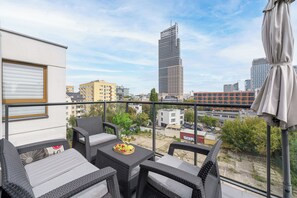 A balcony with wicker furniture, a table with fruit and wine glasses, and a city view including a tall building. - Lumina Comfort 3-Room Apt with Balcony (Warszawa)