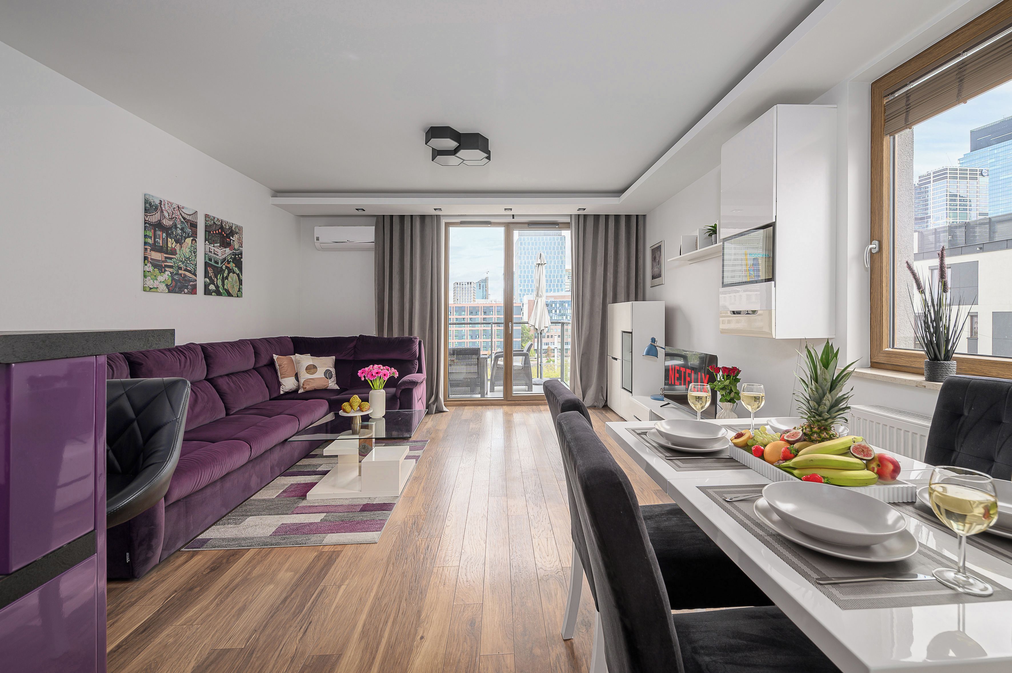 Ein modernes Wohnzimmer mit einem violetten Sofa, einem Essbereich mit Tisch für zwei Personen und Blick auf die Stadt durch große Fenster.
