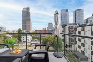 A balcony with wicker furniture, a fruit bowl, and a cityscape view with modern buildings. - Lumina Comfort 3-Room Apt with Balcony (Warszawa)