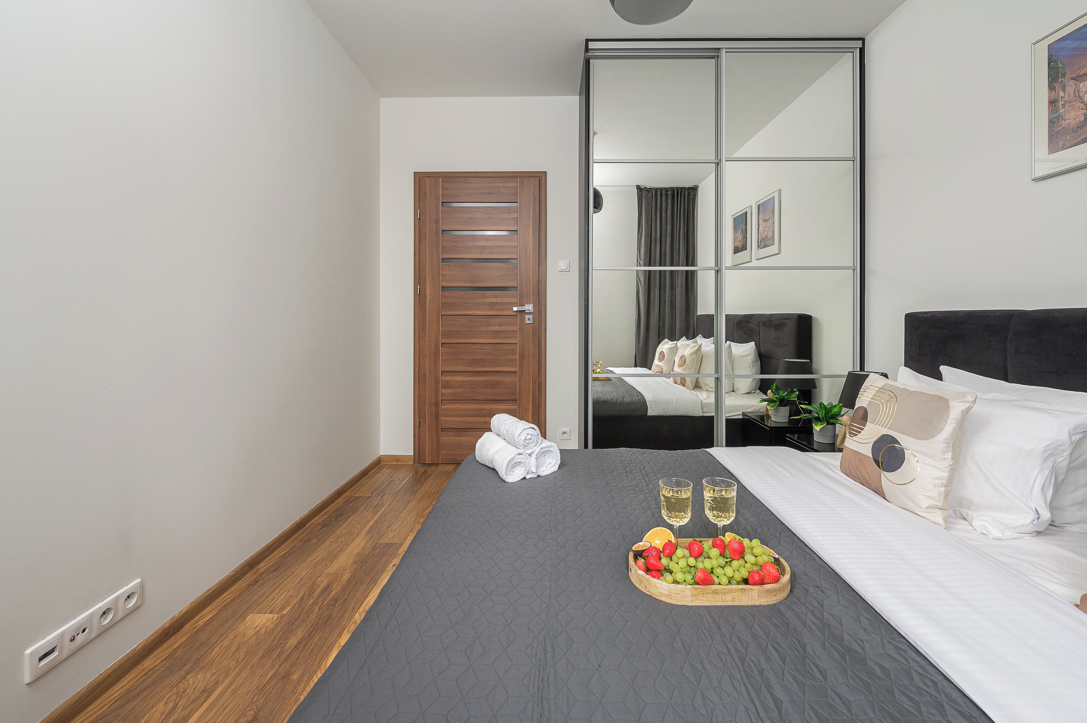 A hotel room with a bed, a wooden door, a mirror, and a tray of fruit and glasses.
