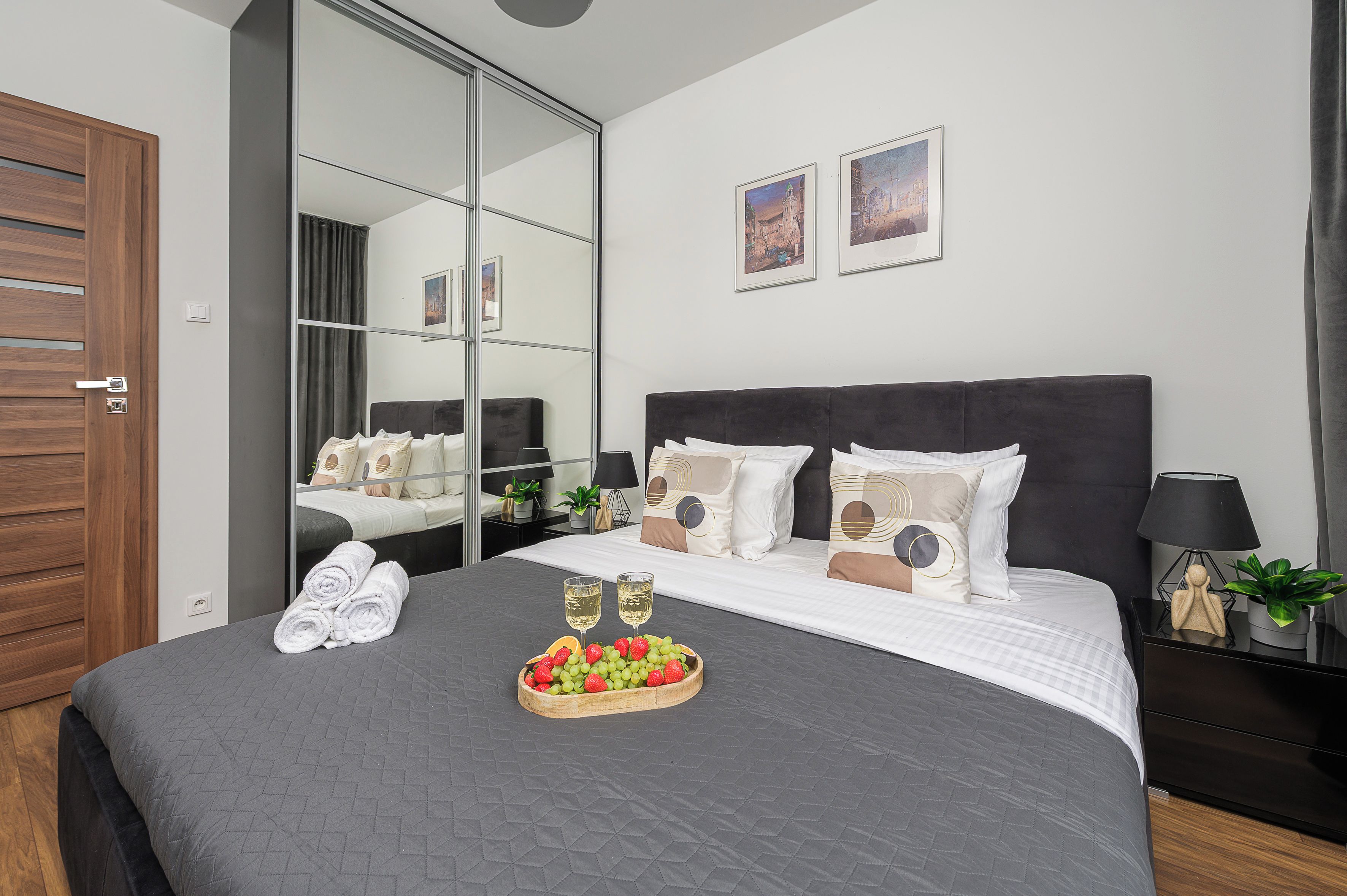 A hotel room with a large bed, a mirror, a nightstand with a lamp, and a tray of fruit and champagne glasses.