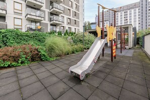 A playground with a slide and climbing structures, surrounded by a hedge and a building with balconies. - Lumina Comfort 3-Room Apt with Balcony (Warszawa)