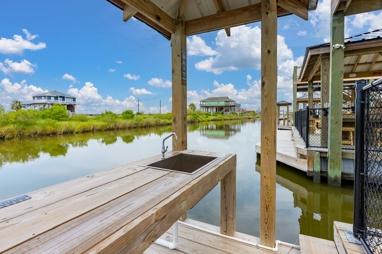 A wooden dock with a sink, a covered area, and a view of a house across a calm body of water.