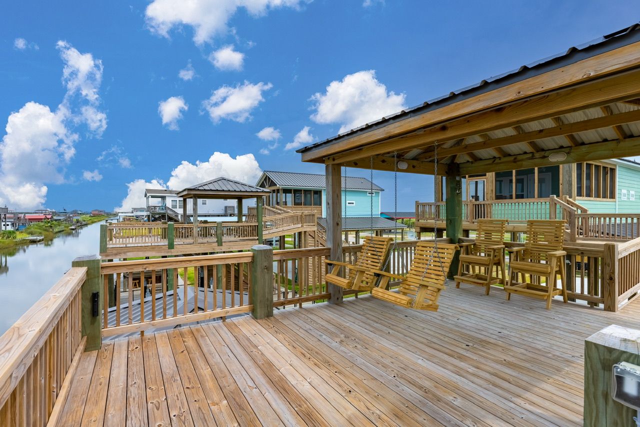 Une terrasse en bois dotée d’un espace salon couvert, offrant une vue sur une étendue d’eau et reliée à des bâtiments par une jetée.