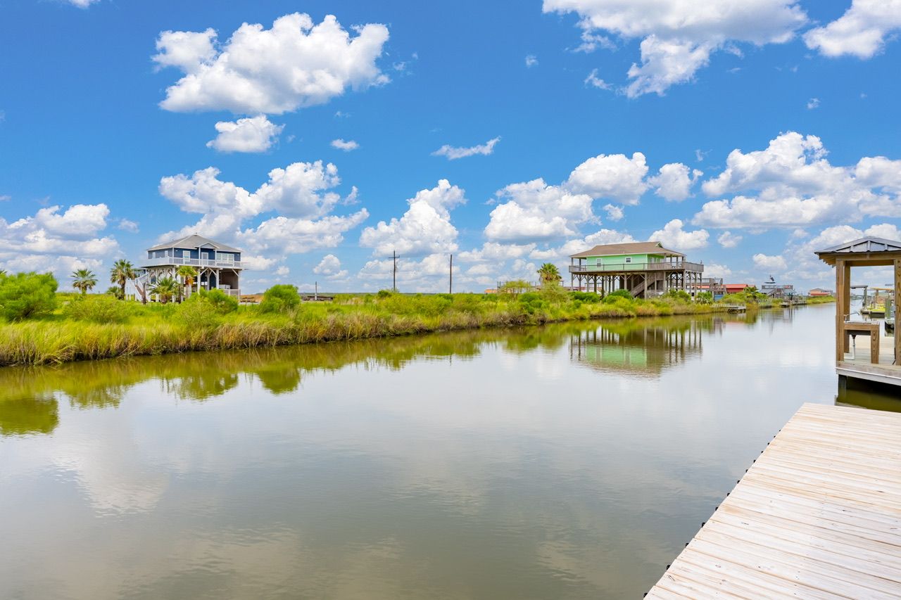 A calm river with wooden docks, two elevated structures with green roofs, and a clear blue sky with clouds.