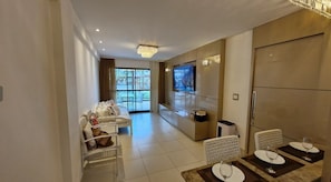 A modern hotel room with a sofa, dining table, TV, and a view of the outdoors. - Muro Alto - Samoa Polynesia - Porto de Galinhas - Ground floor apartment with private pool. (N. Sra. do Ó)