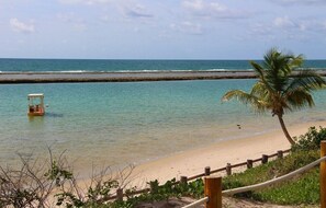 A beach with a wooden fence, a palm tree, and a small boat in the water. - Muro Alto - Samoa Polynesia - Porto de Galinhas - Ground floor apartment with private pool. (N. Sra. do Ó)