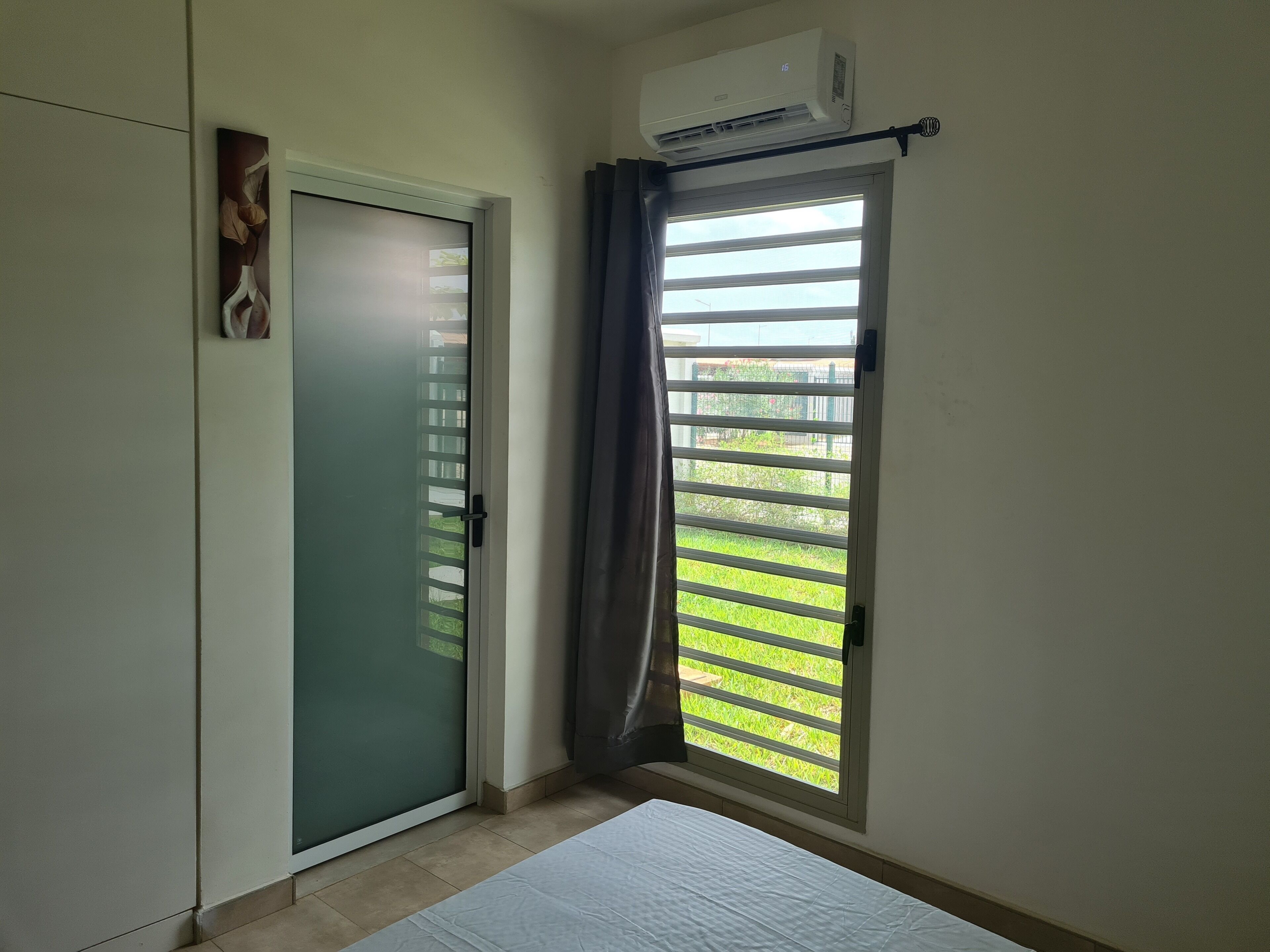 Une pièce dotée d’une porte-fenêtre coulissante donnant sur un balcon, d’un climatiseur blanc fixé au mur et d’un lit avec un matelas blanc.