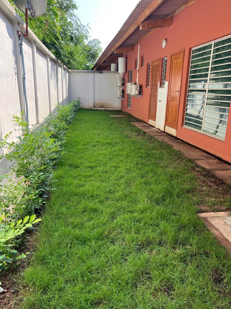 Área gramada bem cuidada com um edifício de fachada vermelha e uma janela com persianas horizontais.
