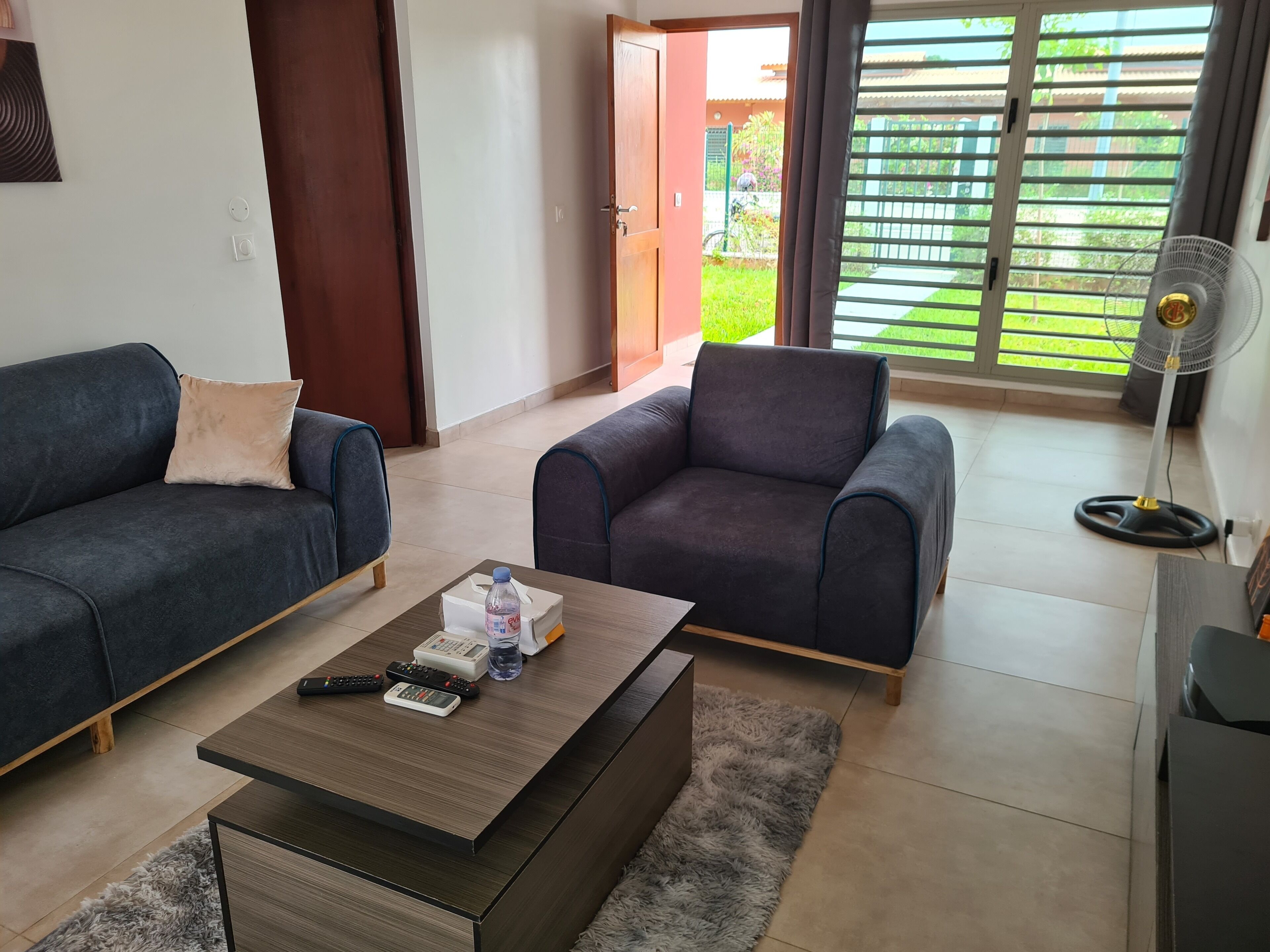 Una sala de estar con dos sofás, una mesa de centro con controles remotos, un ventilador y vista a un jardín a través de una puerta corredera.
