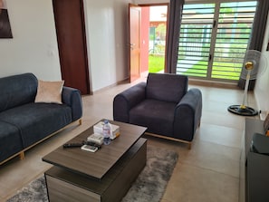 Un salon avec deux canapés, une table basse avec des télécommandes, un ventilateur et une vue sur un jardin, accessible par une porte coulissante.