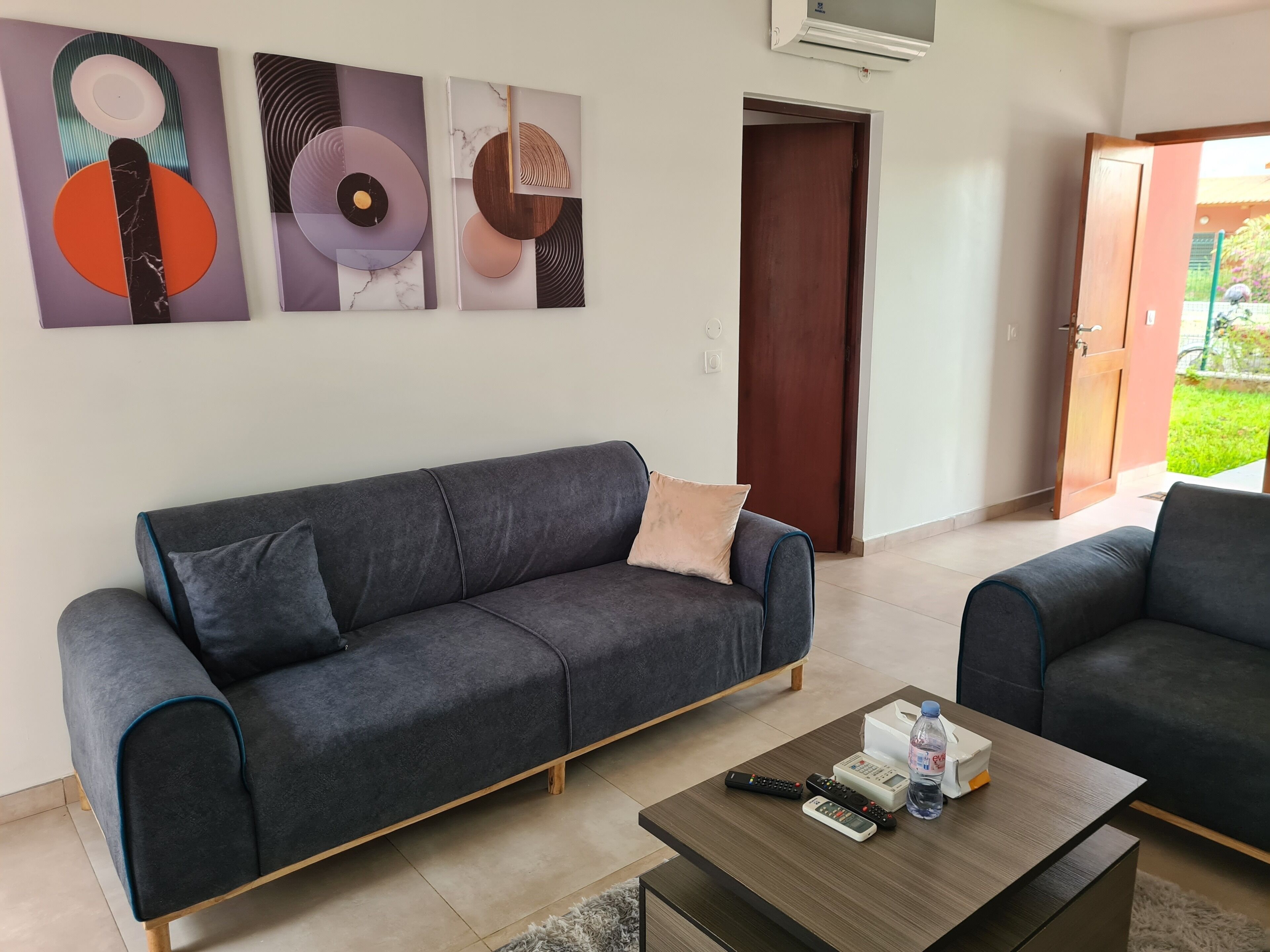 Sala de estar com sofá cinza, mesa de centro com controles remotos e uma garrafa, e arte abstrata na parede.