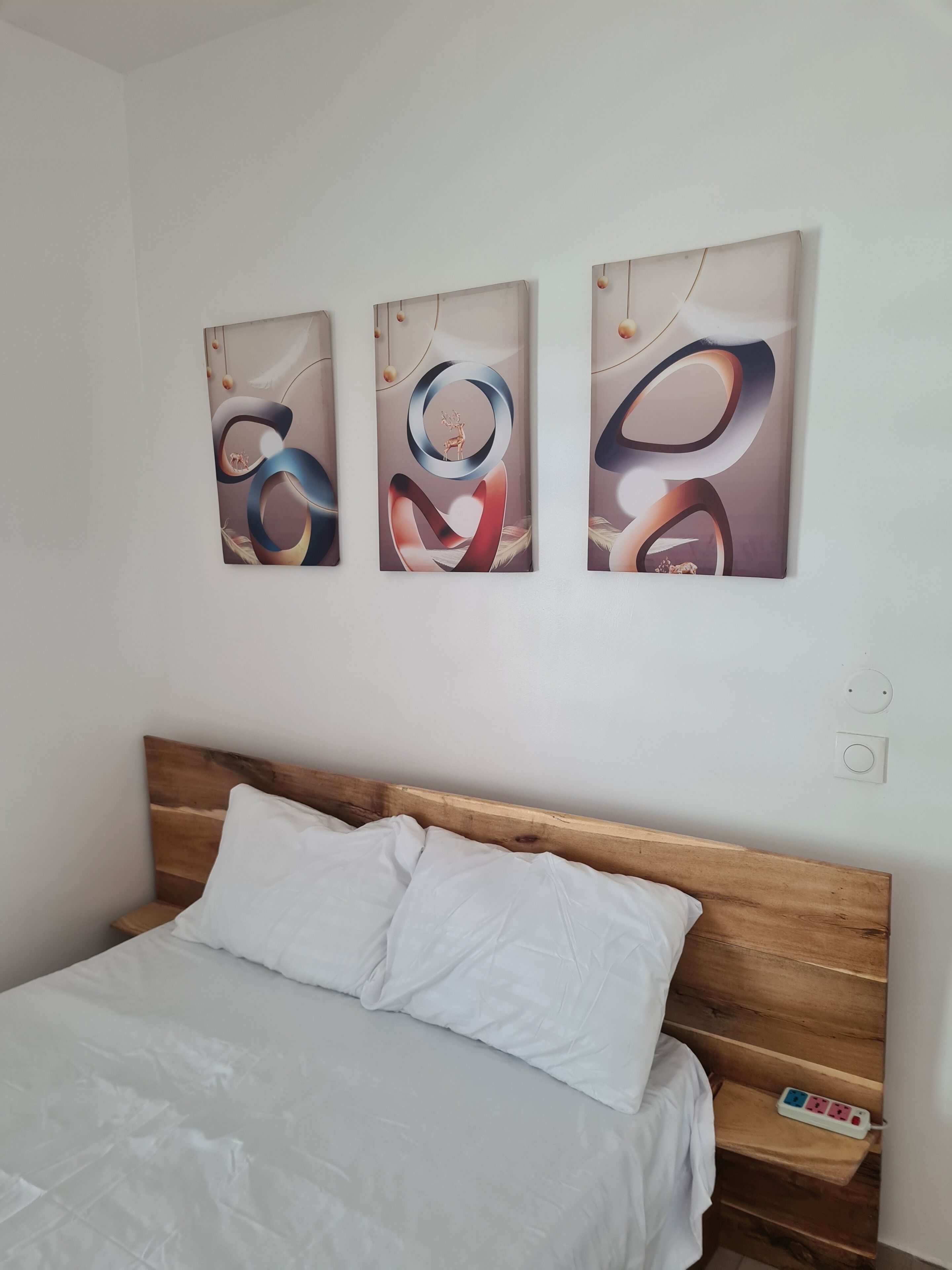 A bedroom with a wooden headboard, a bed with white bedding, and three abstract wall art pieces.