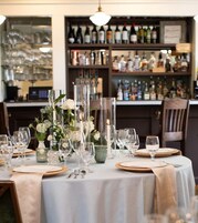Mesa lista para cenar con mantel blanco, cristalería transparente y un centro de mesa con flores y velas.