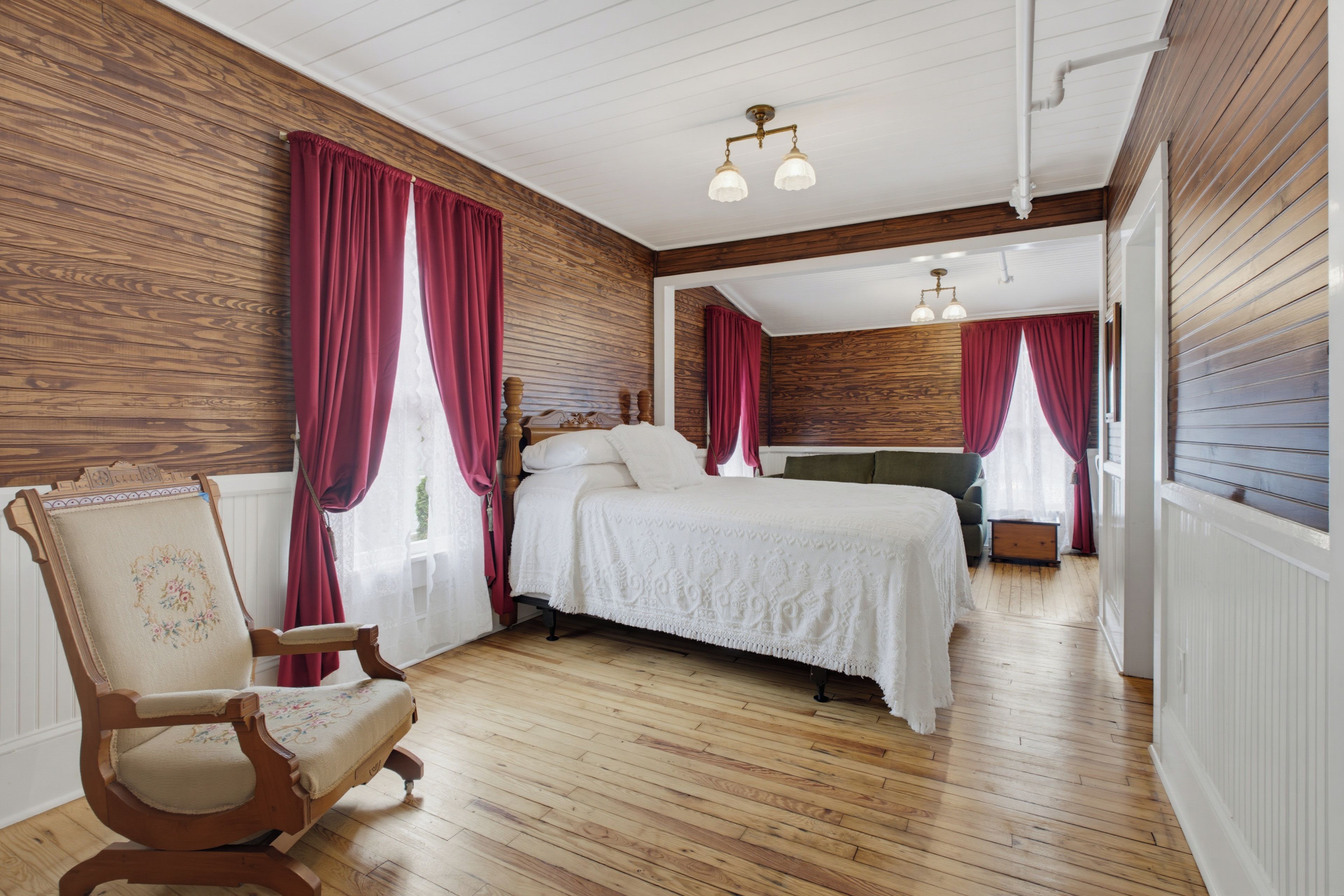 Quarto com piso de madeira, cama com roupa de cama branca, uma cadeira e um sofá. Há duas janelas com cortinas vermelhas.