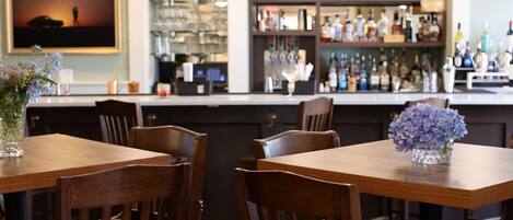 A restaurant interior with wooden tables and chairs, a bar area with shelves of bottles, and a framed picture on the wall.