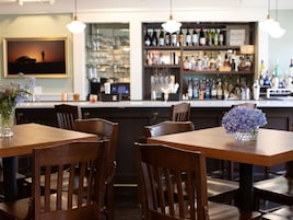A restaurant interior with wooden tables and chairs, a bar area with shelves of bottles, and a framed picture on the wall.