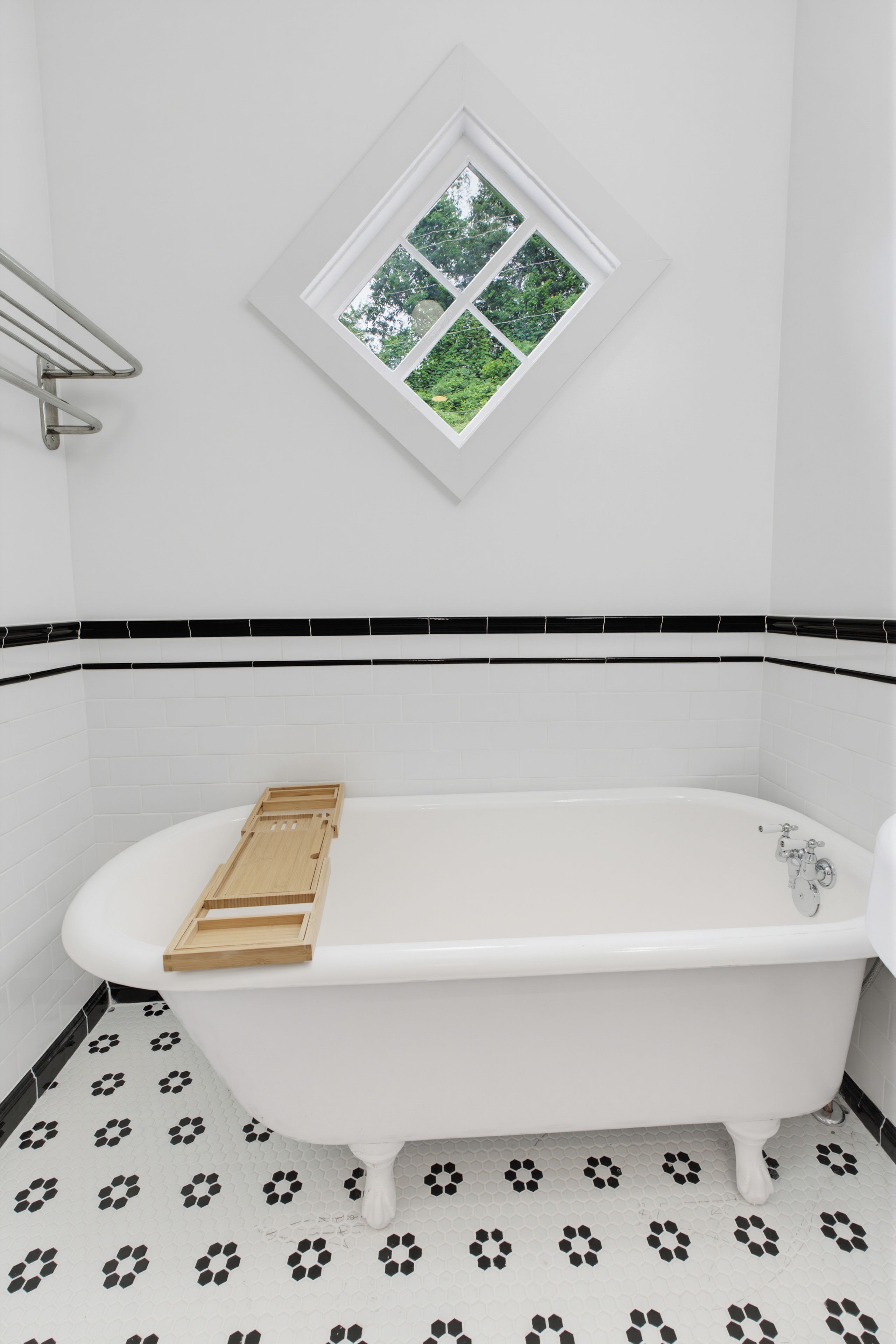 A white freestanding bathtub with a wooden tray, a window with a view of greenery, and black and white patterned floor tiles.